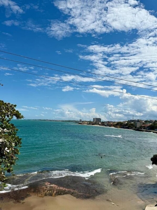A coastal view with clear skies, turquoise water, and a rocky shoreline. - Suítes Prime (Marataízes)