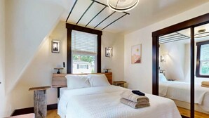 Quarto de hotel moderno com uma cama grande, uma mesa de cabeceira de madeira, uma pequena mesa e uma janela ampla com persianas.