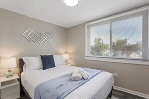 A neatly arranged bedroom with a bed, two bedside tables with lamps, a window with blinds, and a geometric wall decoration.
