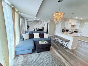 A modern living room with a blue sectional sofa, a small ottoman, and a kitchen area with white cabinets and a wooden countertop.