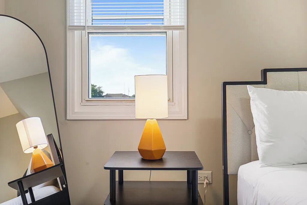 Quarto de hotel com uma cama, mesa de cabeceira com abajur, janela com persianas e espelho fixado na parede.