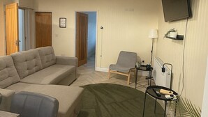Un salon comprenant un canapé d’angle, un tapis rond, un téléviseur fixé au mur et une petite table avec une lampe.