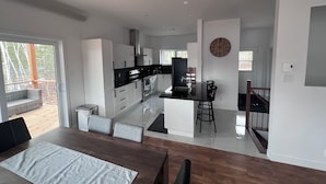 A modern kitchen with white cabinets, a black countertop, and stainless steel appliances. There is a dining area with a wooden table and chairs, and a view of a deck with outdoor furniture. - Chalet 14 pers | Motoneige & Nature | Près du Valinouët - Monts Valin (Saint-David-de-Falardeau)