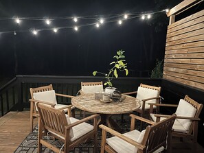 Een terras buiten met een ronde houten tafel, omringd door zes houten stoelen met witte kussens. Boven de tafel hangen lichtslingers.