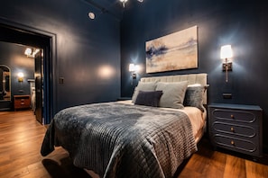 A bedroom with a large bed, a nightstand, a wall-mounted lamp, and a painting. - Hollyhock Suite 401: 4 bed/2 bath with soaking tub (Louisville)