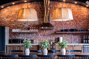 Une cuisine avec un mur en briques, deux suspensions lumineuses, une cuisinière et un coin repas avec des chaises et des vases.