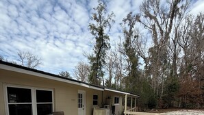 Una terraza techada al aire libre con piso de madera, dos sillas marrones, una mesita y un edificio con ventanas y una puerta.