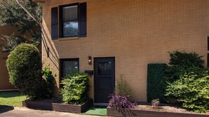 Un immeuble de deux étages en briques, avec une porte d’entrée noire, un petit jardin devant, agrémenté de différents arbustes, et une échelle de secours visible sur le côté gauche.