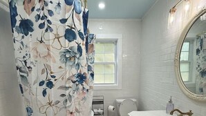 A bathroom with a white bathtub, a toilet, a sink with a mirror, and a window. - The Cottage by Raven`s Keep (Jackson)