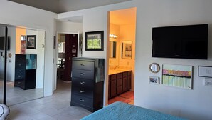 Une chambre à coucher avec un lit, une commode, un téléviseur et une salle de bain visible à travers une porte ouverte.