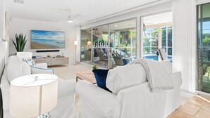 A modern living room with a white sofa, a coffee table, and a TV. There is a glass door leading to a pool area. - Intervillas Florida - Villa Barracuda (Cape Coral)