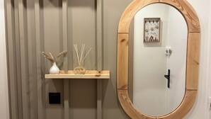 Un bagno con uno specchio ovale con cornice in legno, uno scaffale in legno con oggetti decorativi e un interruttore della luce fissato al muro.