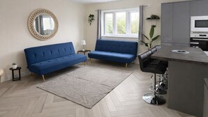 Un salon moderne avec deux canapés, un parquet à motifs chevrons, un coin cuisine et une grande fenêtre.