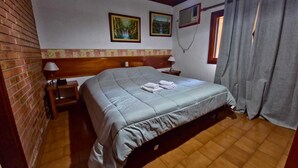 Ein Schlafzimmer mit Holz-Kopfteil, einem Bett mit grauer Bettdecke, zwei Bildern an der Wand, einem Fenster mit Vorhängen und einer Ziegelwand.