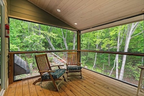 A wooden deck with two chairs and a large window overlooking a forest. - Mountain Escape w/ Hot Tub + Game Room Sleeps 6 Near Lake Chatuge (Hiawassee)