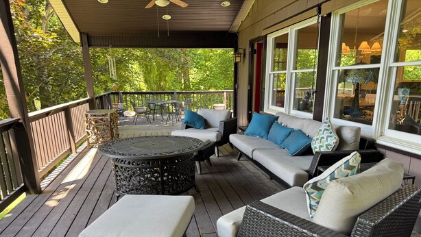 A covered outdoor deck with wicker furniture, a round table, and a view of the surrounding greenery. - Experience Stunning Sunsets & Mountain Views in a Cozy Retreat near Asheville (Fairview)