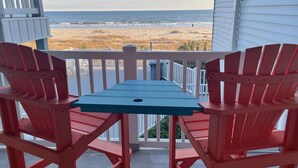 A balcony with red Adirondack chairs and a blue table, overlooking a beach and ocean. - Oceanfront and Steps Away from the Pool and Beach at Ocean Isle Beach, NC! (Ocean Isle Beach)