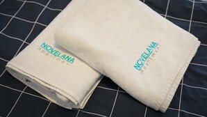 Two folded white towels with 'NOVELANA' embroidered on them, placed on a dark blue bed with a grid pattern. - Novelana (Banja Luka)