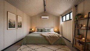 A bedroom with a bed, two bedside tables with lamps, a window with curtains, a wall-mounted air conditioner, and a shelf with decorative items. - Tiny Home (Casula)