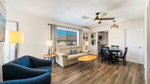 Ein Wohnzimmer mit Sofa, Couchtisch, Esstisch und Stühlen. Es gibt einen Deckenventilator, ein Fenster mit Jalousien und eine Tür.