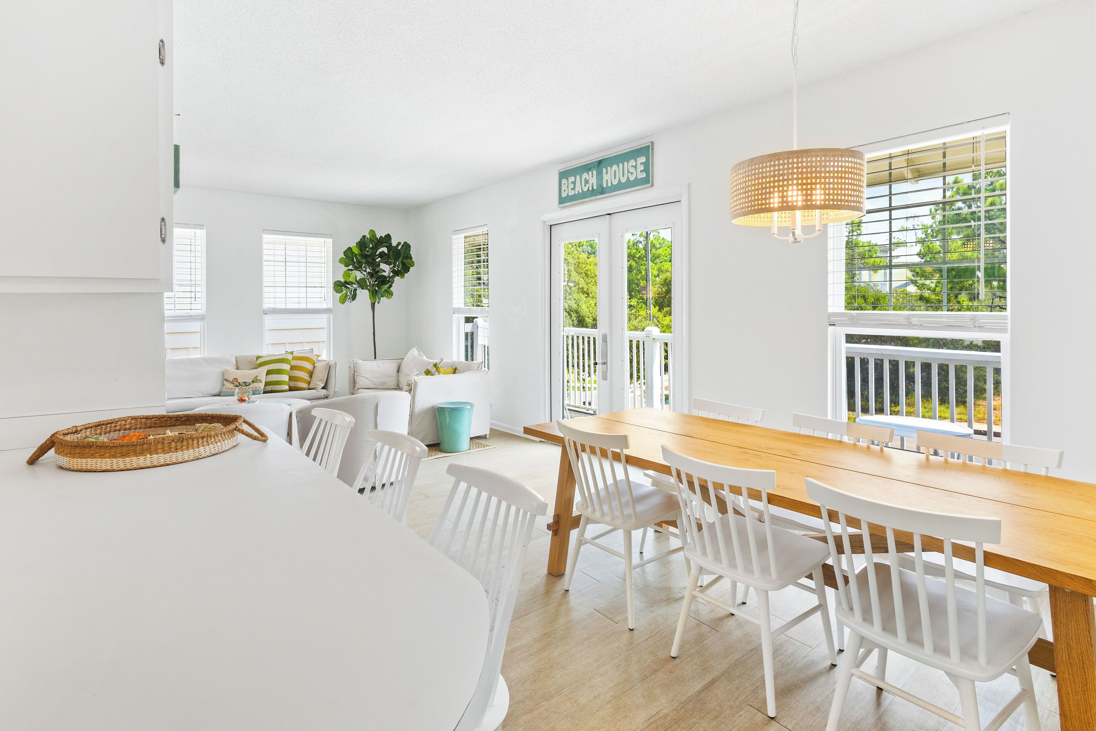 A bright, white interior with a dining area, a sign reading "BEACH HOUSE," and a view of greenery outside.