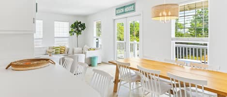 A bright, white interior with a dining area, a sign reading "BEACH HOUSE," and a view of greenery outside.