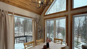 A dining area with a wooden table and chairs, large windows, and a chandelier. - The Beaver Den • Snowmobile & Ski Escape (Dysart Et Al)
