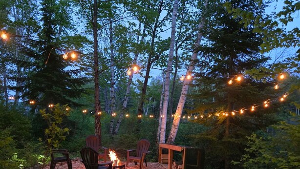 An outdoor seating area with wooden chairs and a fire pit, surrounded by trees and string lights. - The Beaver Den • Snowmobile & Ski Escape (Dysart Et Al)