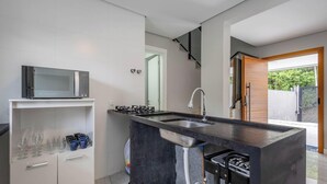 A modern kitchen with a built-in microwave, stainless steel sink, and a gas stove. There is a storage cabinet with wine glasses and cups, and a view of the outdoors through a glass door. - House w/ Terrace & BBQ 850m From the Beach Jsf0748 (Bombinhas)