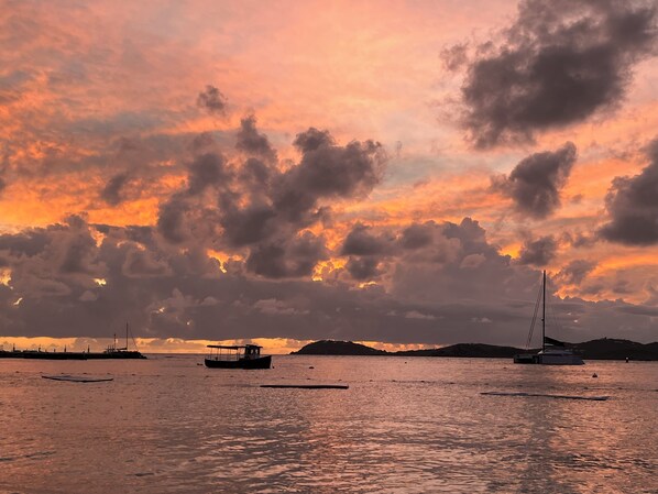 Pôr do sol sobre um mar calmo, com barcos e terra ao longe.