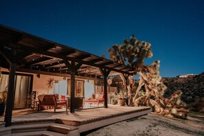 A covered outdoor area with wooden furniture and a large cactus. - Retreat on an acre! Near Joshua Tree Nat. Park, stunning views, pool, hot tub! (Yucca Valley)