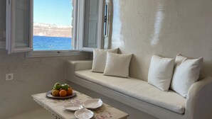 Una habitación con un sofá blanco, una mesita con frutas y vajilla, y vista al mar a través de una ventana.