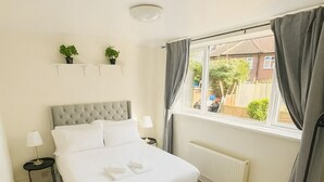 Quarto com cama, mesas de cabeceira, radiador, janela com cortinas e vista para o exterior.