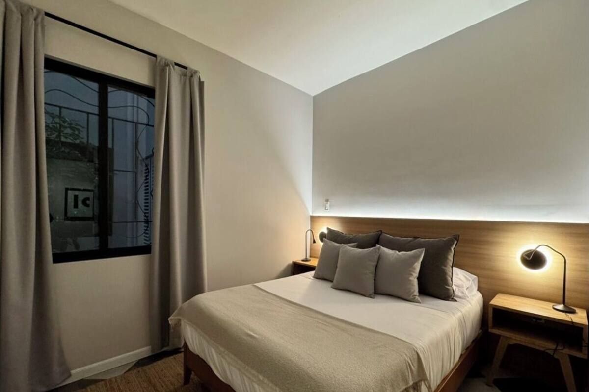 Un dormitorio con cama, mesitas de noche, lámpara, ventana con cortinas y vistas al exterior.