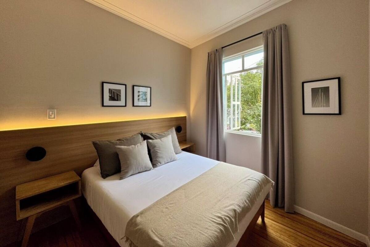 Un dormitorio con cama, mesita de noche, ventana con cortinas y cuadros en la pared.