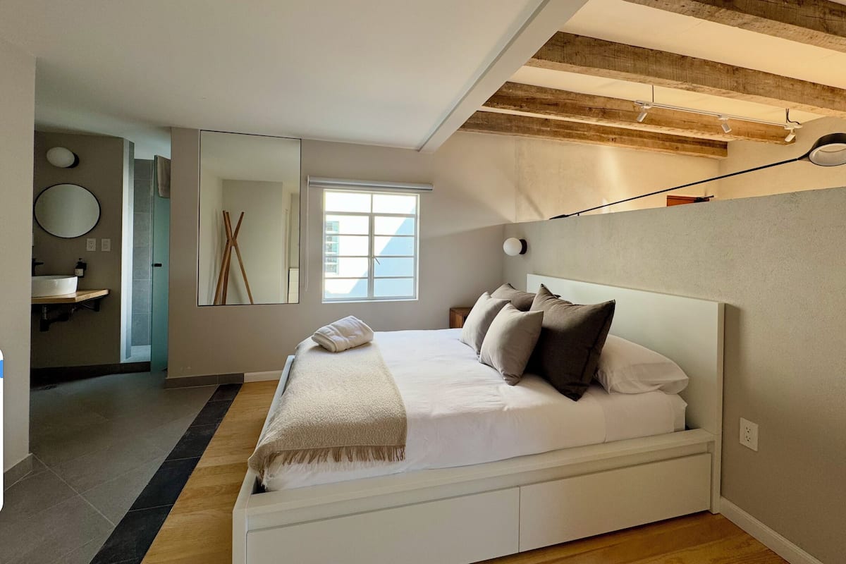 Un dormitorio con cama, ventana, espejo y baño con lavamanos.
