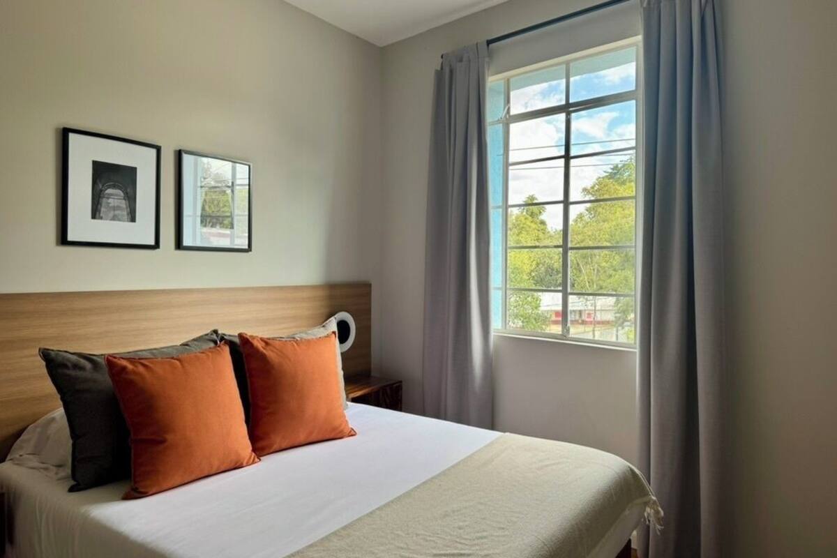 Habitación de hotel con una cama, dos cuadros en la pared, una ventana con cortinas y vistas a zonas verdes.