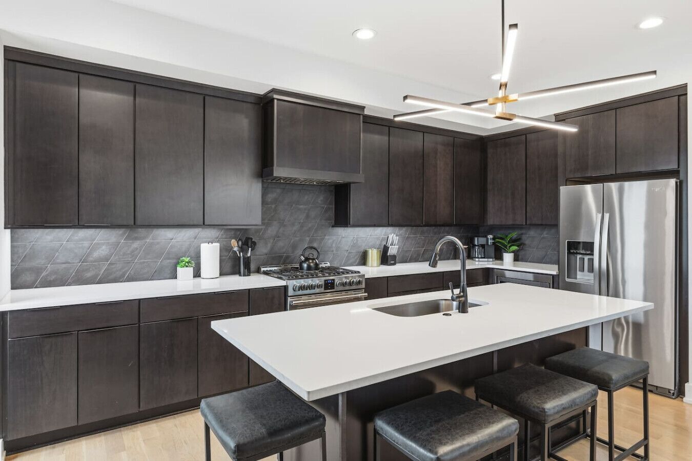 Una cocina moderna con armarios oscuros, encimera blanca, una isla central con taburetes, un refrigerador de acero inoxidable y un ventilador de techo.