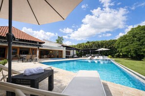 A swimming pool with lounge chairs and umbrellas, surrounded by a house with a tiled roof and a garden. - Exclusive 5-bedroom villa in Casa de Campo (La Romana)