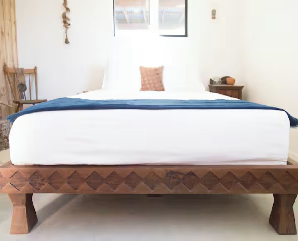Uma cama com colchão branco e manta azul, uma cabeceira de madeira com padrão de losangos e uma pequena mesa de cabeceira com abajur.