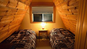 A small attic bedroom with two beds, a window with blinds, a table with a lamp, and a fan. - Cozy 2-bedroom chalet in lovely Carrabassett Valley (Carrabassett Valley)