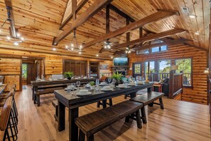 Un vaste espace dédié à la salle à manger, avec des tables et des bancs en bois, une cheminée en pierre et de grandes fenêtres offrant une vue sur l’extérieur.