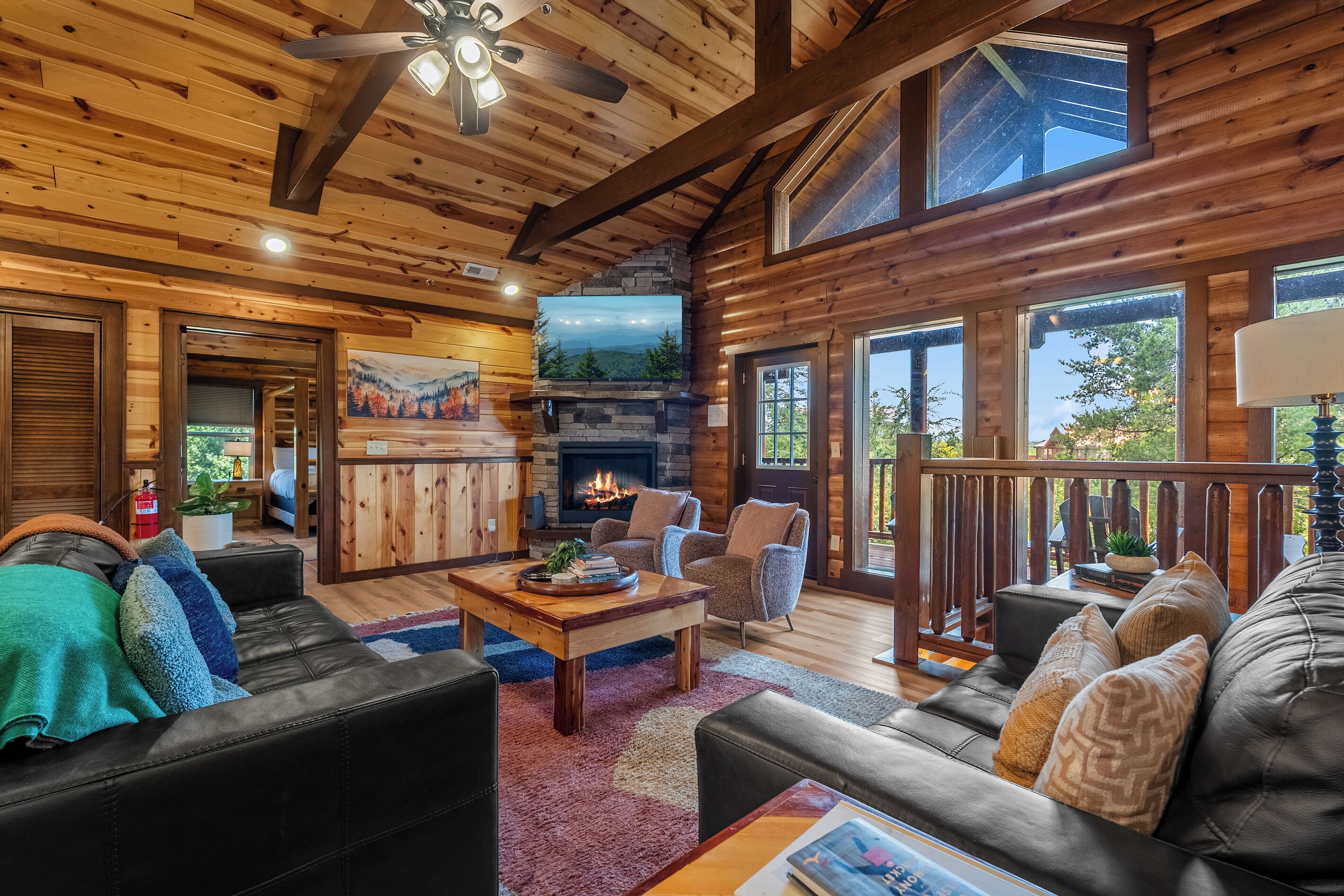 A cozy log cabin interior with a fireplace, a ceiling fan, and a large window overlooking a forested area.