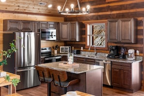 A modern kitchen with wooden paneling, stainless steel appliances, granite countertops, and a central island with bar stools. - 8BR Luxury Lodge w/ Indoor Pool, Gazebo & Mountain Views Sleeps 28 (Sevierville)