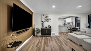 A modern living room with a flat-screen TV, a black cabinet, a dining area, and a kitchen in the background. - Welcome to Chic Parma House, a stylish 5-bedroom, 1-bath home with boutique-inspired design, perfect for families, traveling professionals, and extended stays just minutes from Cleveland, hospitals, and major highways. (Parma)