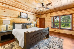A wooden cabin bedroom with a bed, nightstands, a ceiling fan, and a large window with a view of greenery. - 6BR Lodge in the Smokies w/ Indoor Pool, Arcade & Views Sleeps 22 (Sevierville)