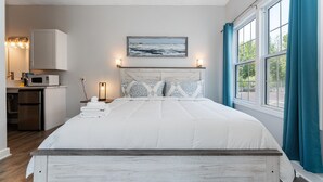 Une chambre à coucher comprenant un lit, une tête de lit, un tableau, une table de chevet, une lampe et une fenêtre avec des rideaux.