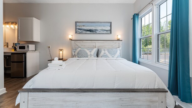 A bedroom with a bed, a headboard, a painting, a nightstand, a lamp, and a window with curtains.
