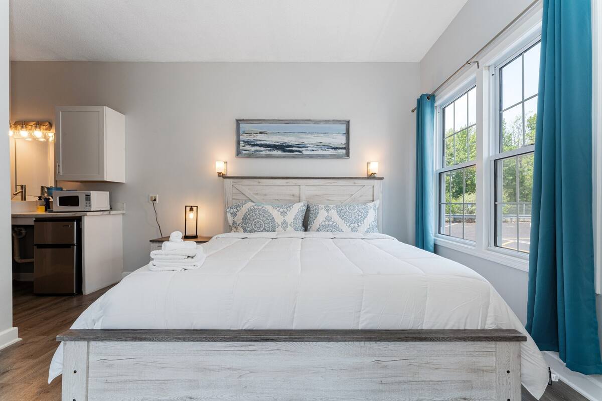 A bedroom with a bed, a headboard, a painting, a nightstand, a lamp, and a window with curtains.