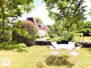 A garden with a white table and chairs, a stone wall, and a house with a red roof in the background. - Gîtes de France® - La Longère de Bessous (CONCORES)